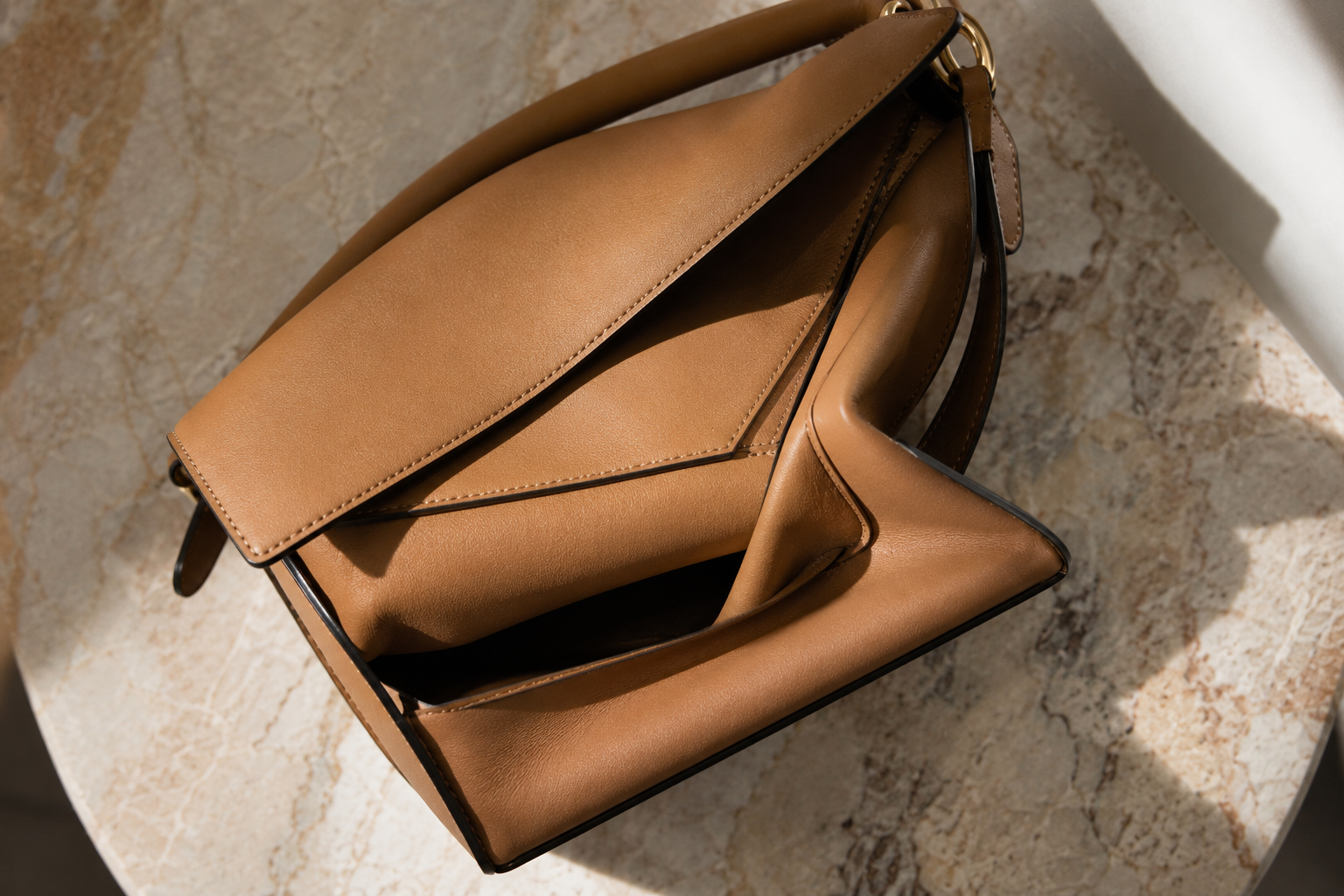 Overhead close-up of a tan Loewe Puzzle bag partially collapsed on a marble table showing the folded geometric leather panels and embossed Anagram