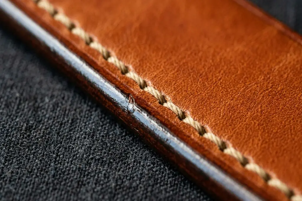 Close up of a cognac leather belt edge showing burnished finishing and neat saddle stitching under raking light