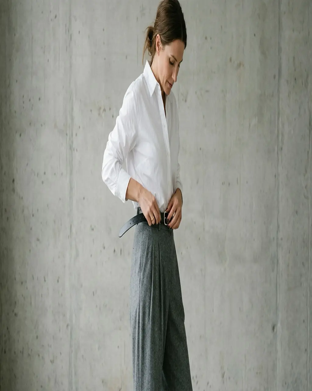 Person wearing pleated grey trousers and a white shirt fastening a slim black leather dress belt at the waist