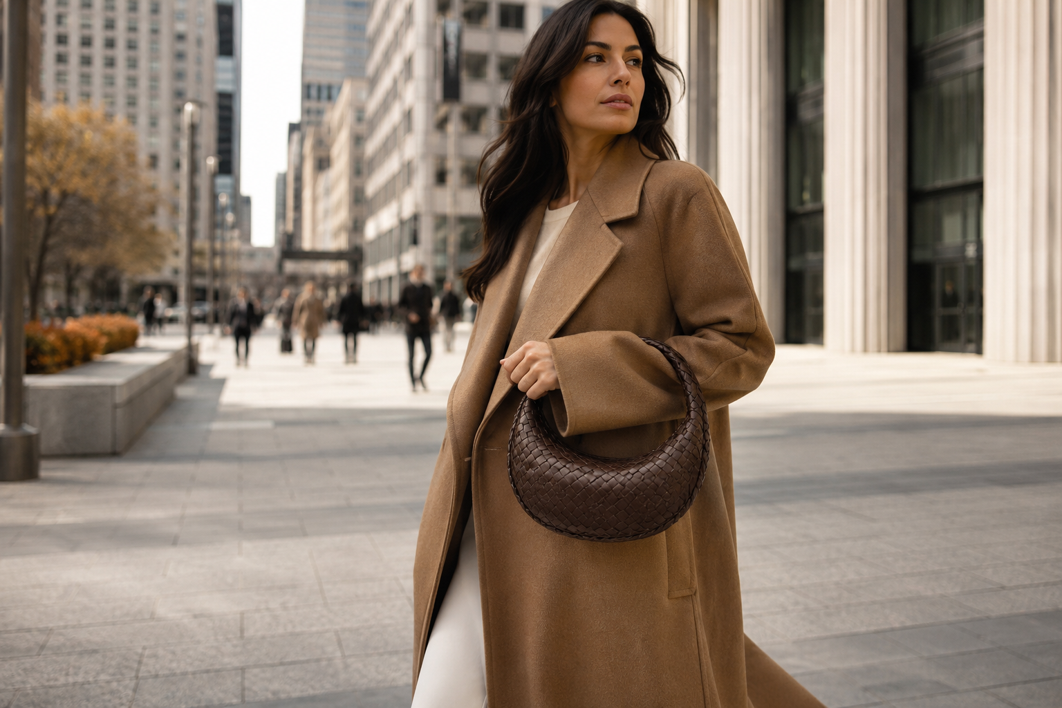 Italian-Canadian woman crossing a sunlit Toronto plaza in a camel wool coat carrying a chocolate-brown Bottega Veneta Intrecciato Jodie bag