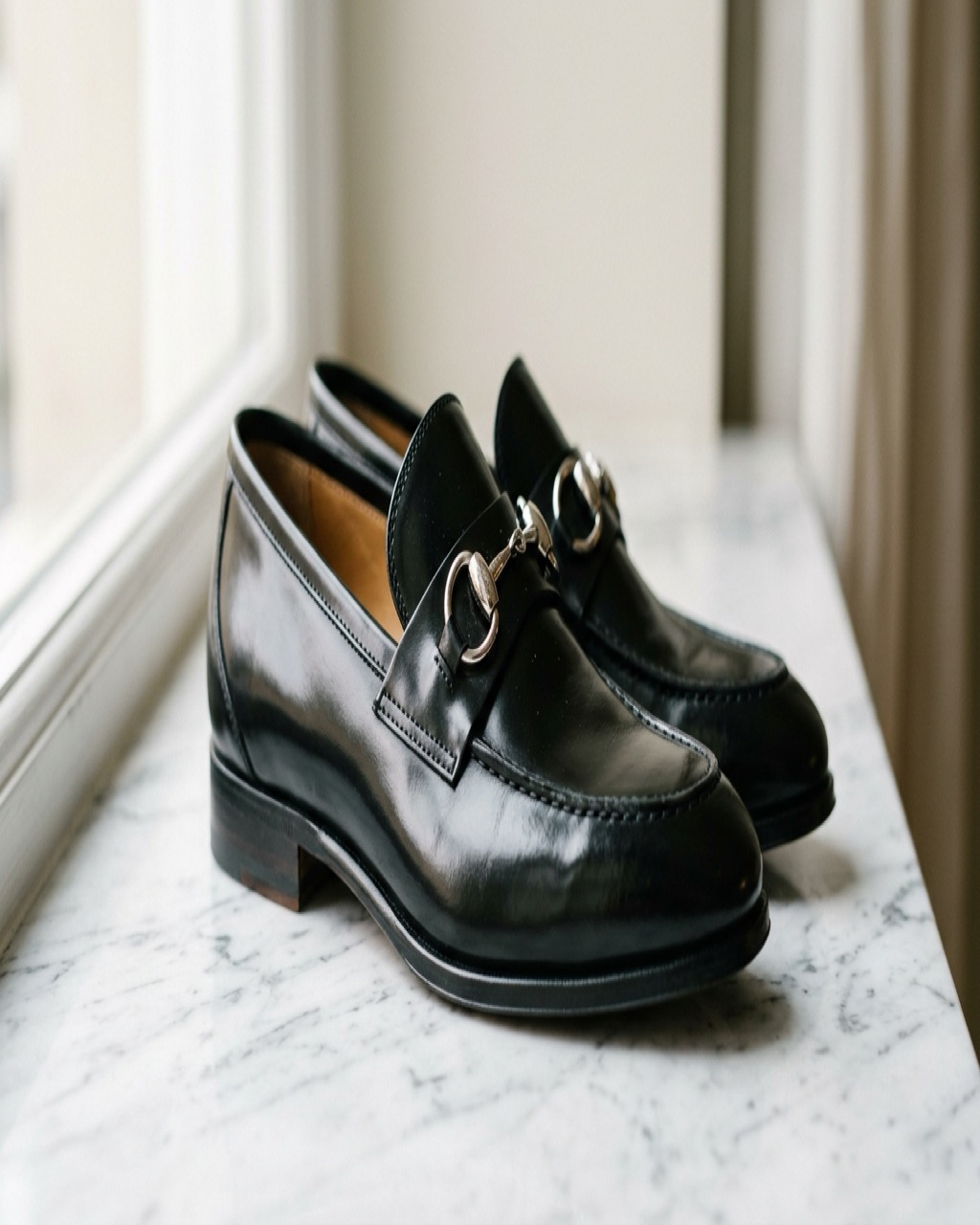 Close-up of black leather loafers with a gold metal bit detail resting on white marble under soft studio lighting in an editorial fashion photograph