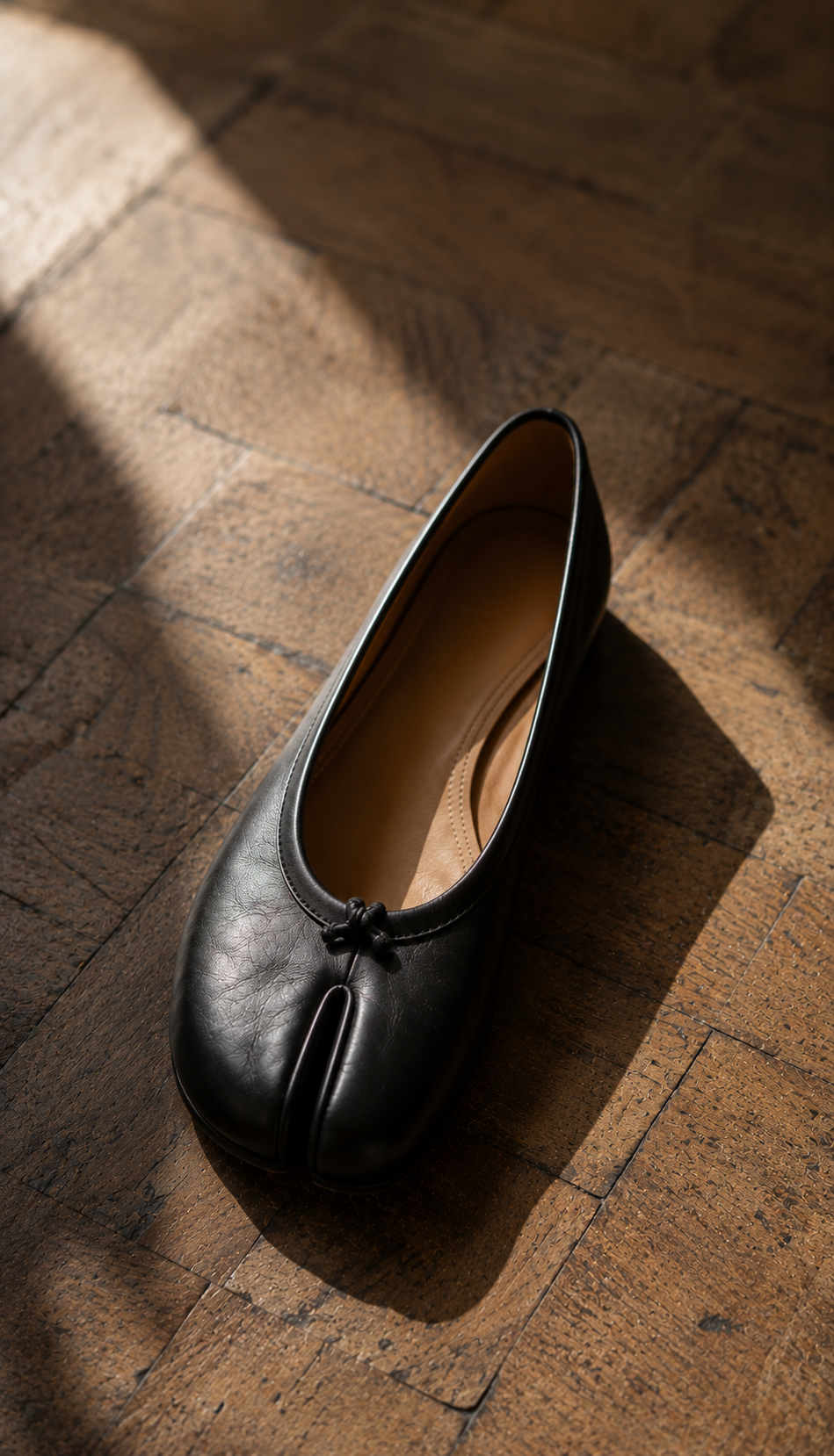 Close-up of a single black leather Tabi ballet flat on a worn parquet floor, split toe and stitched seam clearly visible, soft window light from the left